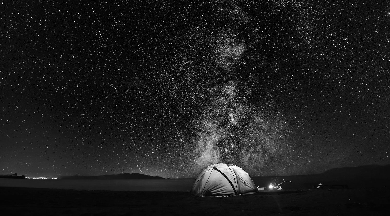 Star-filled night sky over a desert landscape with illuminated tent beneath the Milky Way